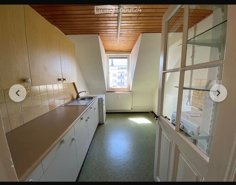 The kitchen features a wooden ceiling, white cabinets, a sink with a faucet, and an oven. The space is well-lit with natural light coming through a window, and the floor is a light green color.