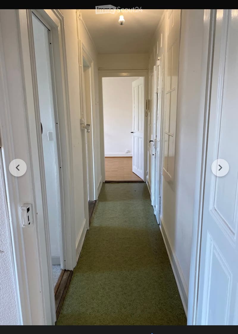 A long, narrow hallway with white walls, green carpeting, and multiple doors on both sides. The hallway leads to an open area with wooden flooring visible at the end.