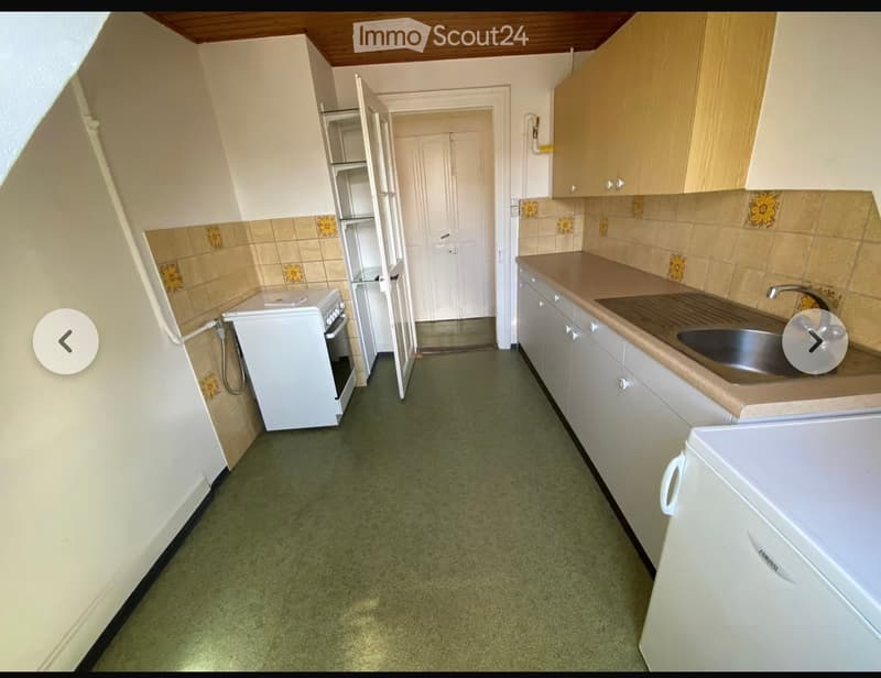 A compact kitchen with white cabinets, a sink, an oven, and a refrigerator. The walls are tiled with floral designs, and the floor is green.