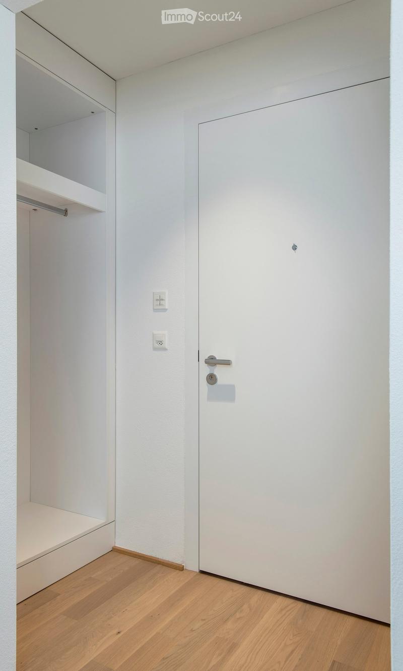 Empty room with white walls, wooden floor, built-in shelving unit, and a closed white door.