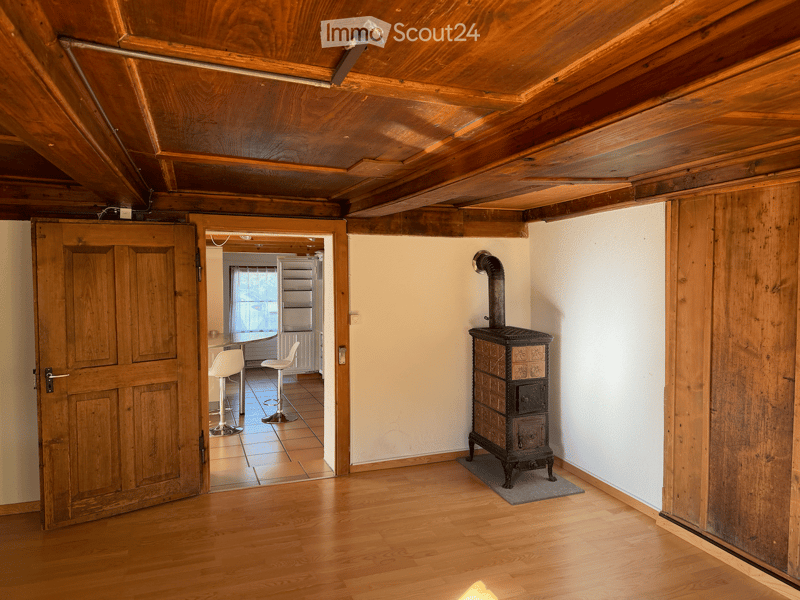 The room features exposed wooden beams on the ceiling, wooden flooring, and a wooden stove. A doorway leads to another room with two white stools and a small table.