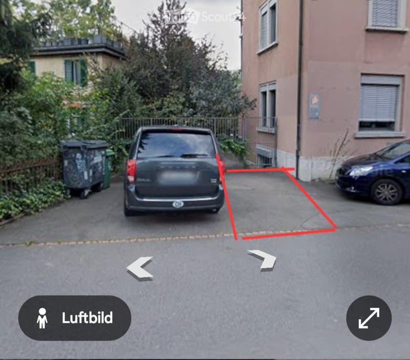 Parking area next to a building with two cars parked, surrounded by greenery and a fence.