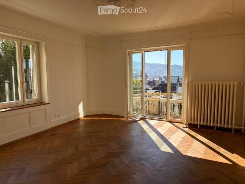 Empty room with wooden herringbone flooring, large windows, and a radiator. The room has a balcony accessible through large glass doors, with a scenic view of mountains and cityscape.