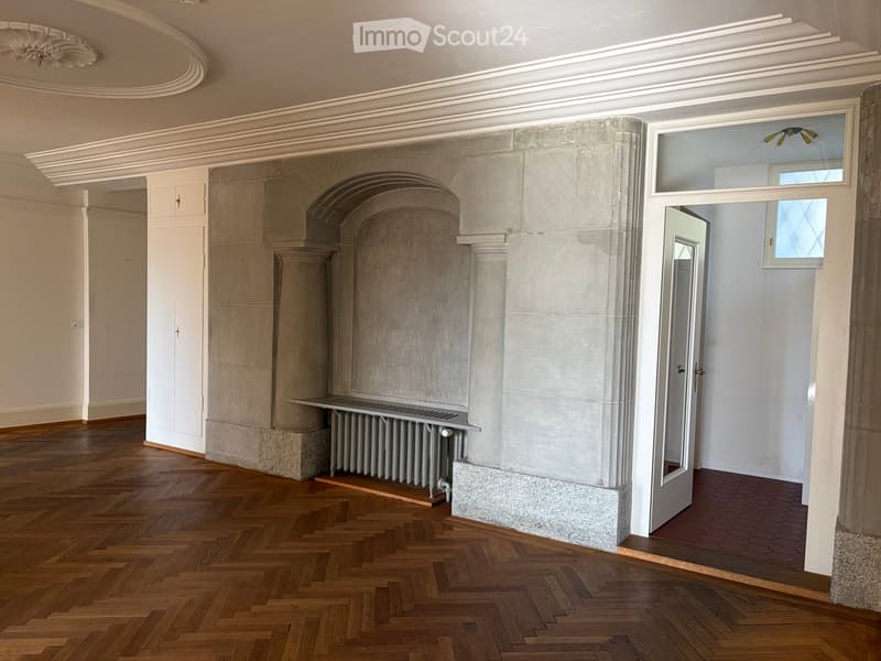 Empty room with wooden herringbone flooring, white walls, and a large stone fireplace with a radiator below. A white cabinet is visible on the left, and a doorway leads to another room with a red brick floor.