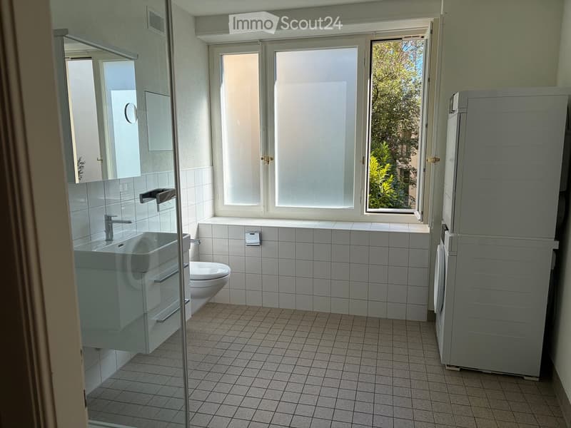 Modern bathroom with white tiled walls and floor, a sink with a chrome faucet, a toilet, a mirrored cabinet, and a large window letting in natural light.