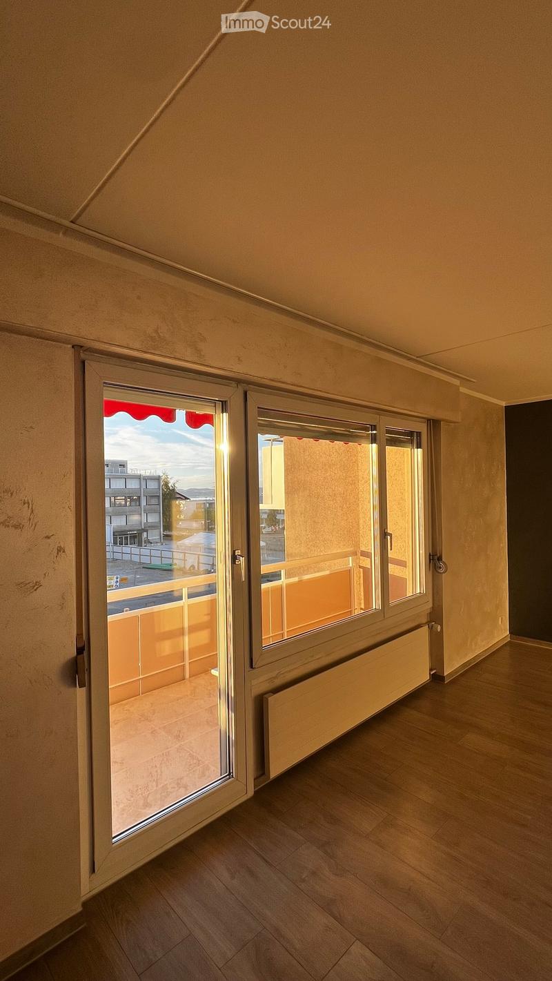 Empty room with wooden flooring, large window with open sliding doors leading to a balcony, radiator visible under the window.