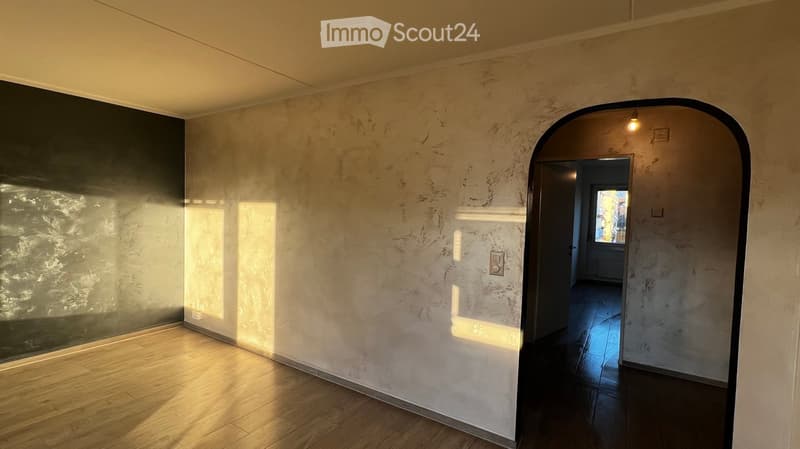 Empty room with wooden flooring, textured walls, and a large arched doorway leading to another space.