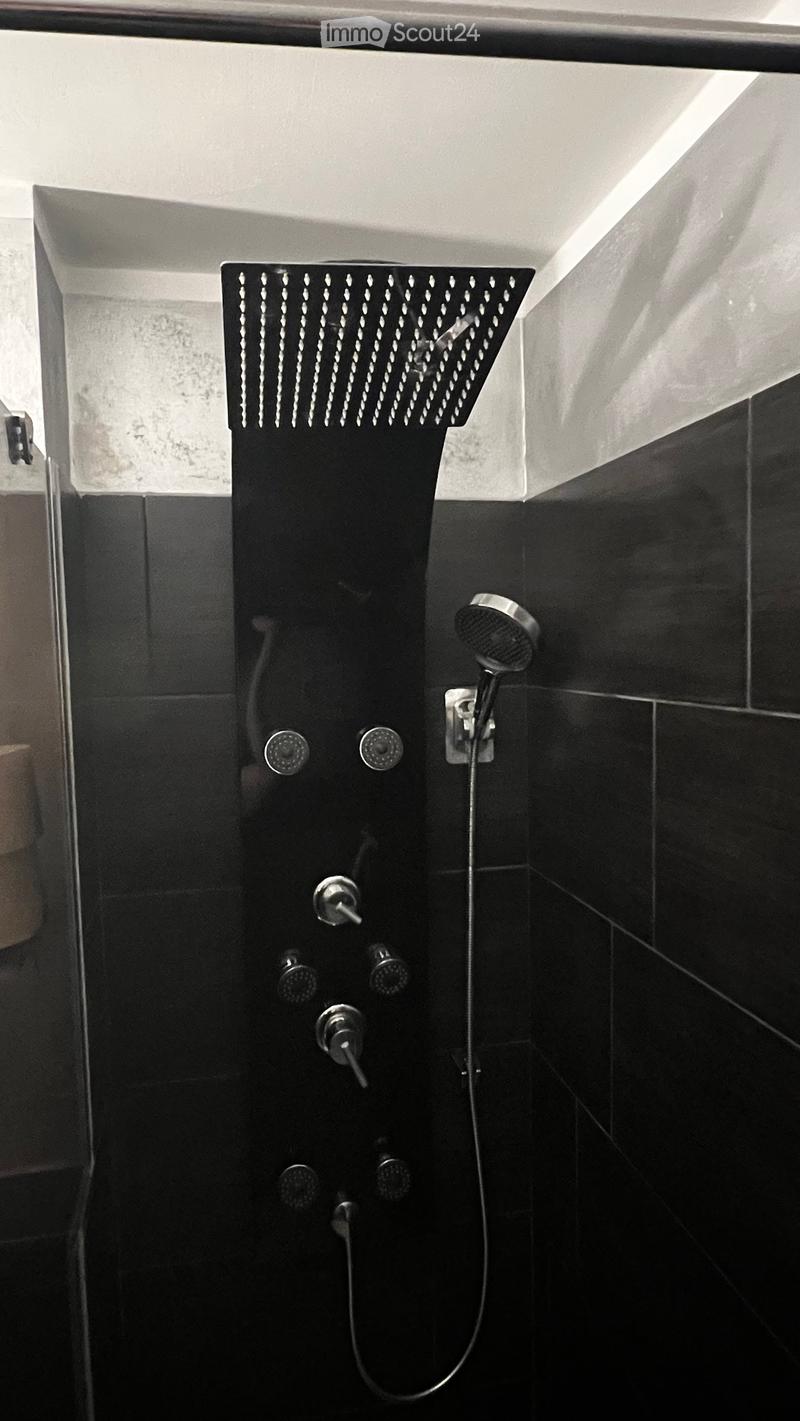 Modern black tiled shower stall with a large, perforated showerhead and multiple control knobs for water flow and temperature.