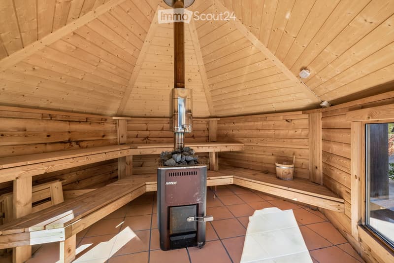 Wooden sauna with benches, a stove, and a bucket, featuring a sloped wooden ceiling and tiled floor.