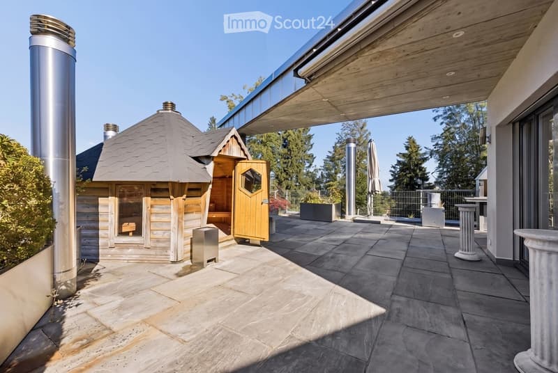 A modern outdoor patio area with a wooden sauna, stone tiles, and a covered section with wooden beams. The area includes a tall chimney, a closed umbrella, and surrounding greenery.
