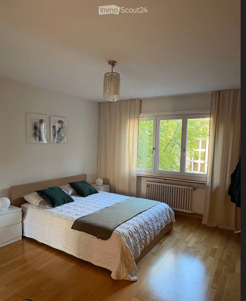 Bright bedroom with wooden flooring, large window with curtains, bed with green pillows and white comforter, nightstand with lamp, and wall-mounted picture frames.