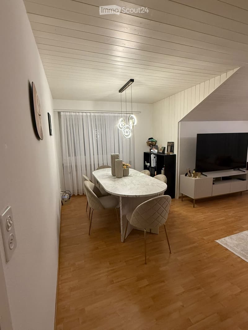 Modern dining area with a white marble dining table, six chairs, a TV stand with a flat-screen TV, a shelf with decorative items, and a hanging light fixture above the table.