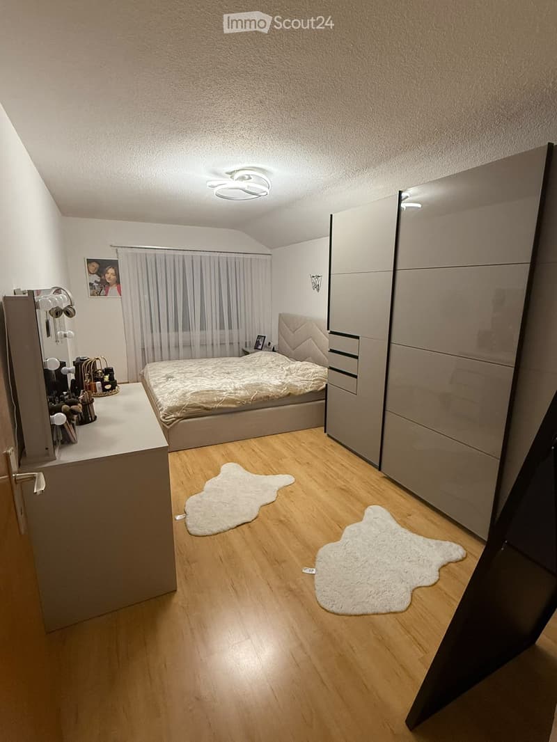 Modern bedroom with wooden flooring, a large bed with a textured headboard, a mirrored wardrobe, a vanity table with mirrors, and two white rugs on the floor.