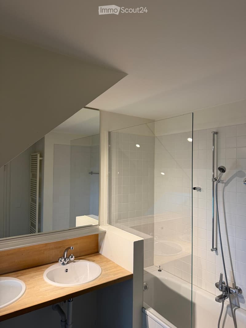 Modern bathroom with a wooden vanity, two sinks, a large mirror, a bathtub with a glass shower enclosure, and a handheld showerhead.