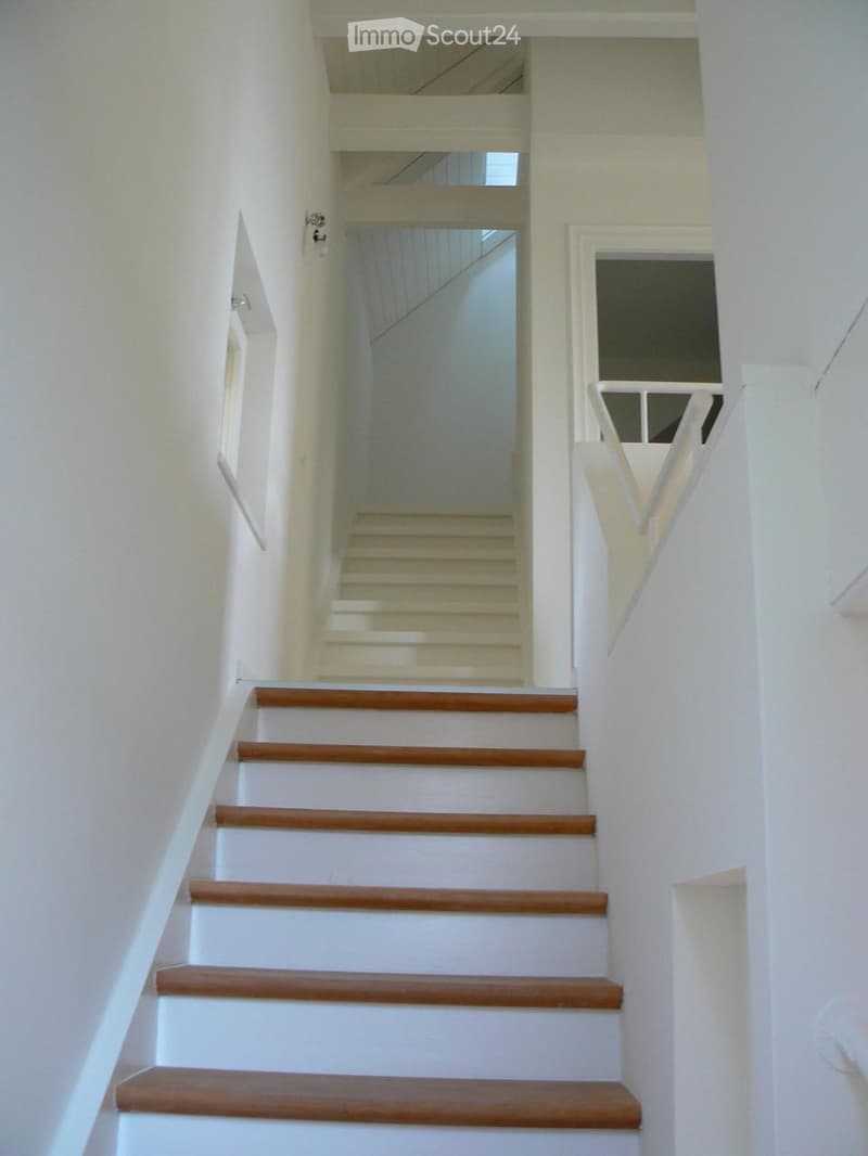 A modern staircase with wooden treads and white risers, featuring white walls and a white handrail, leading to an upper level with a visible window at the top.