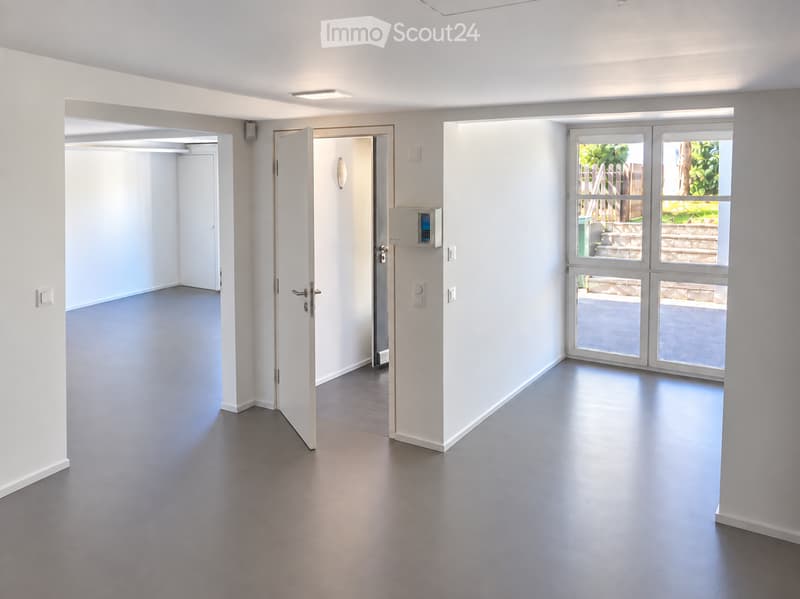 Empty room with white walls, grey flooring, and large windows leading to an outdoor area. No furniture or equipment is present.
