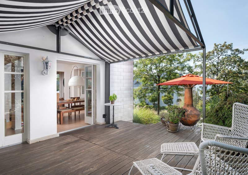 Wooden deck with white wicker chairs, small table, outdoor umbrella, and a large ceramic vase. The deck is covered by a black and white striped awning, with a view of a garden and trees.