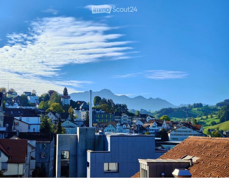 A panoramic view of a town with a mix of modern and traditional buildings, surrounded by green hills and distant mountains under a clear blue sky.