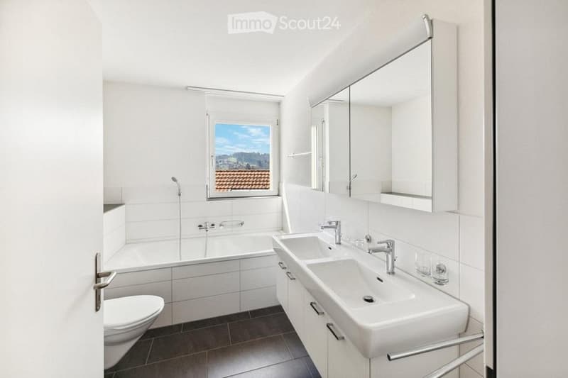 Modern bathroom with white tiles, a bathtub, toilet, double sink, mirrored cabinet, and a window offering a scenic view.
