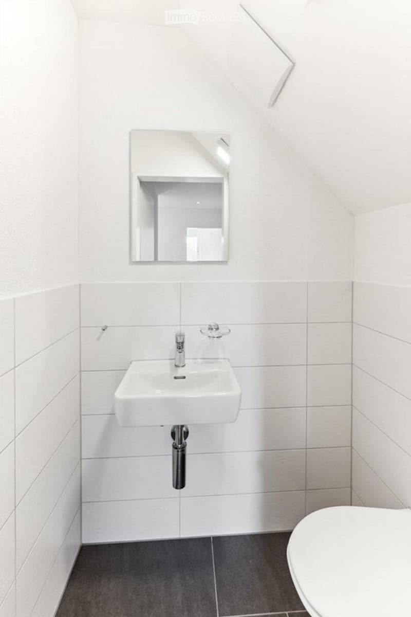 Modern, minimalist bathroom with white tiles, a white sink, a mirror, and a toilet. The walls are white, and the floor is dark grey.