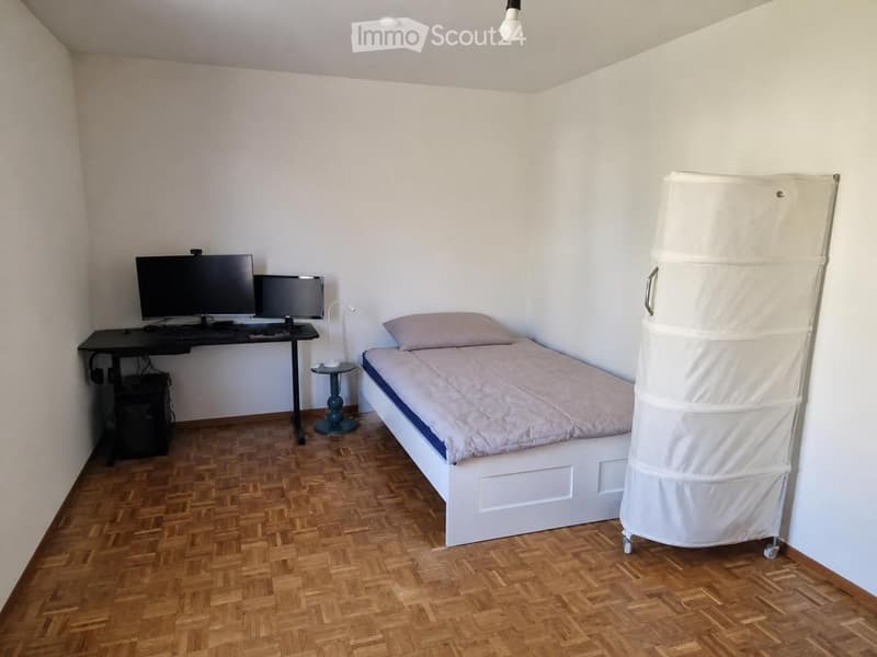 A small, furnished room with a single bed, desk, two screens, a chair, and a lamp. The room features wooden flooring and white walls.