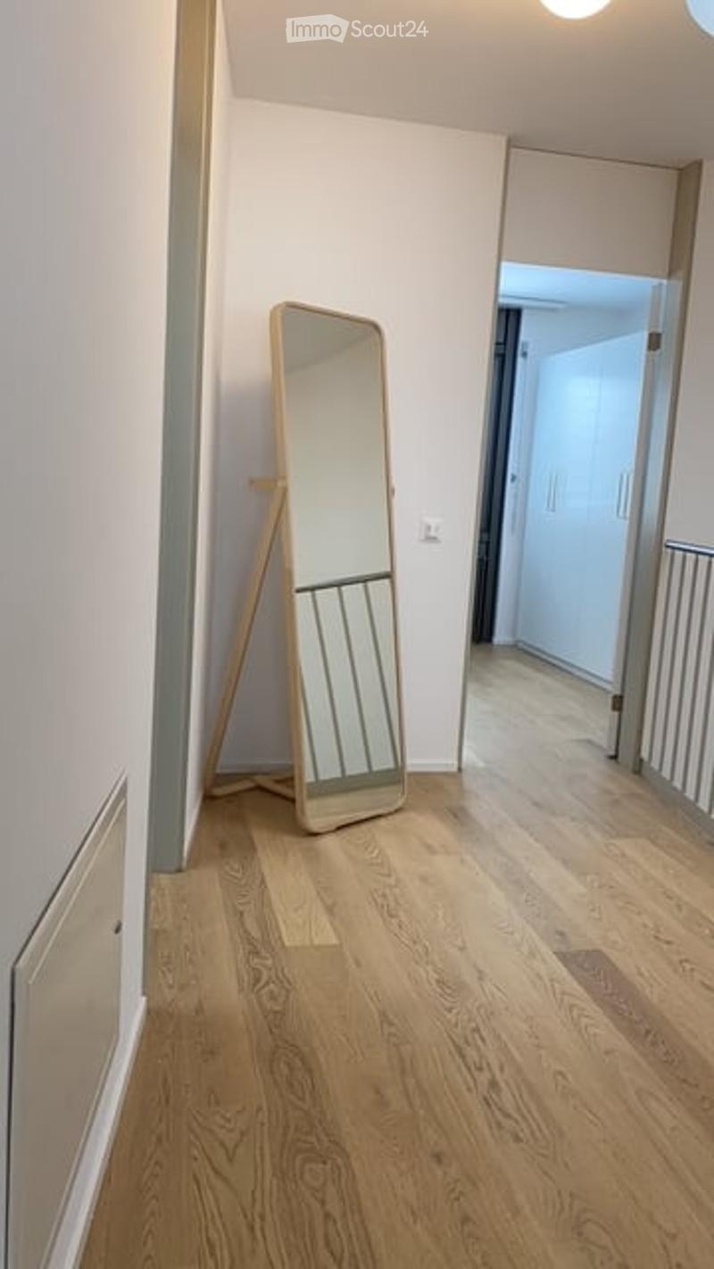 A clean, modern hallway with light wooden flooring, white walls, and a large floor mirror leaning against the wall. A doorway leads to another room, and a small railing is visible on the right side.