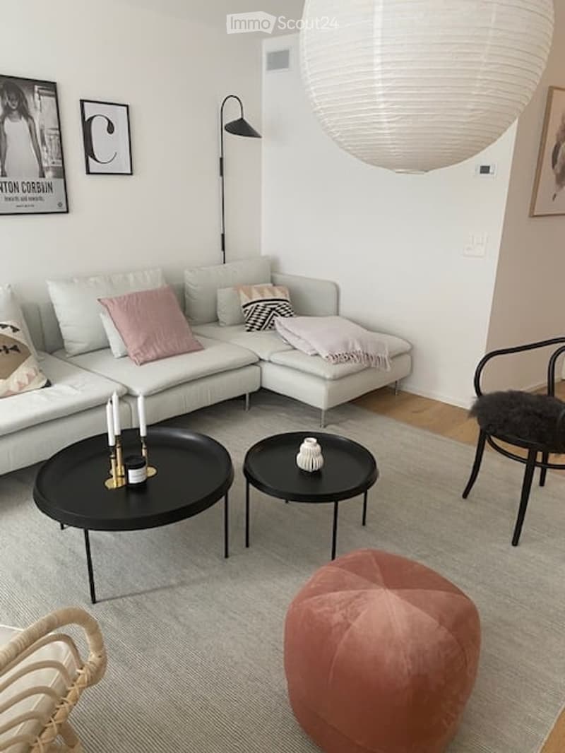 Modern living room with a white sectional sofa, pink and grey throw pillows, two black coffee tables, a floor lamp, a pink ottoman, and a black chair with a fur throw.