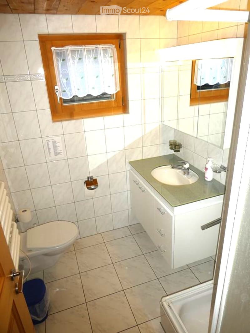 A small bathroom with white tiled walls and floor, featuring a toilet, a sink with a mirror above it, and a window with a white curtain. A soap dispenser and a small cabinet are also visible.