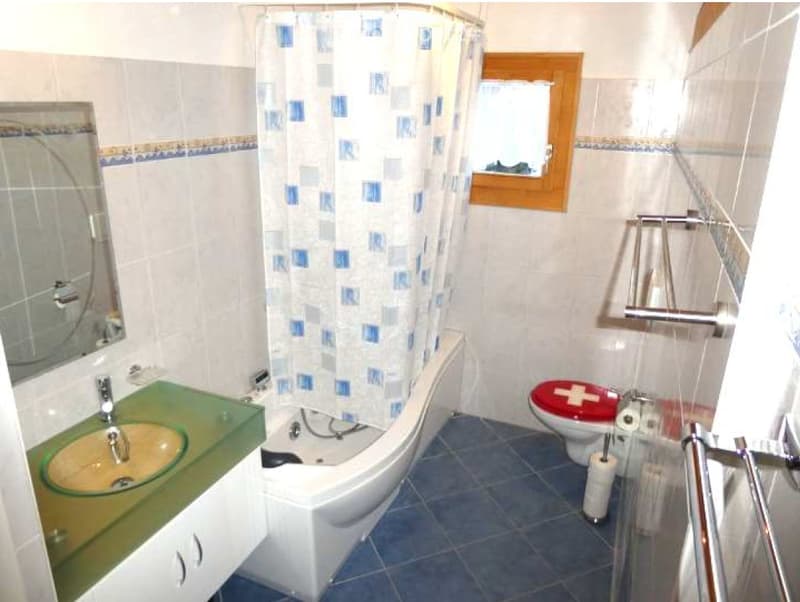 Bathroom with a white bathtub, a sink with a green countertop, a toilet with a red seat cover, a mirror, and a shower curtain with a blue and white pattern.
