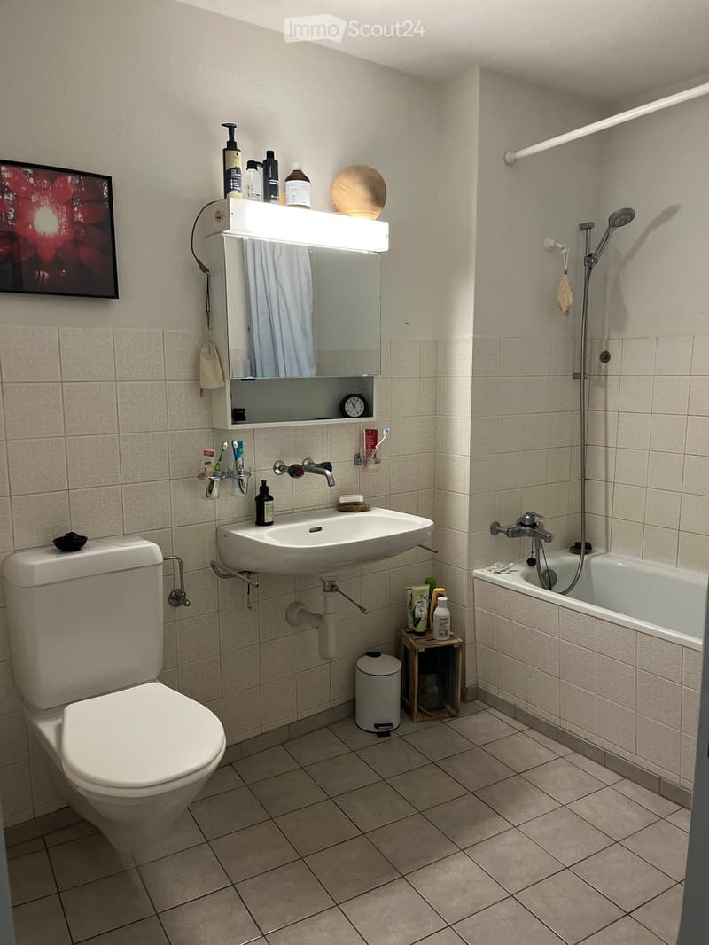 Modern bathroom with beige tiles, a toilet, a sink with a mirror cabinet, a bathtub with a showerhead, and various toiletries.