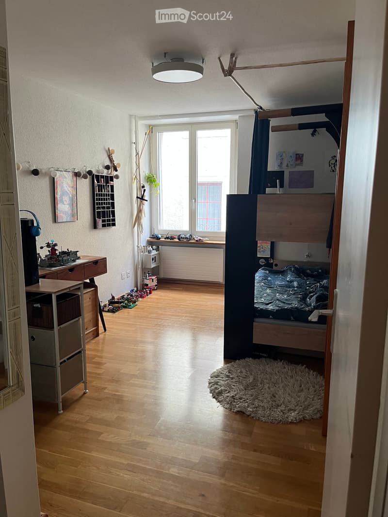 A well-lit bedroom with wooden flooring, a lofted bed, a desk with a chair, a storage unit, a large window with a curtain, and a shaggy rug on the floor.