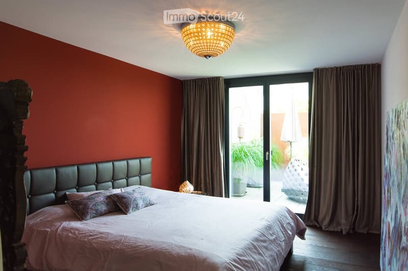Modern bedroom with a large bed, leather headboard, patterned pillows, and a chandelier. Large windows with curtains lead to a balcony with outdoor furniture.