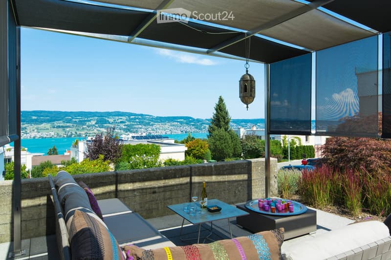 A modern outdoor terrace with a panoramic view of a city and water, featuring a sofa, a small table with wine and glasses, and decorative cups.