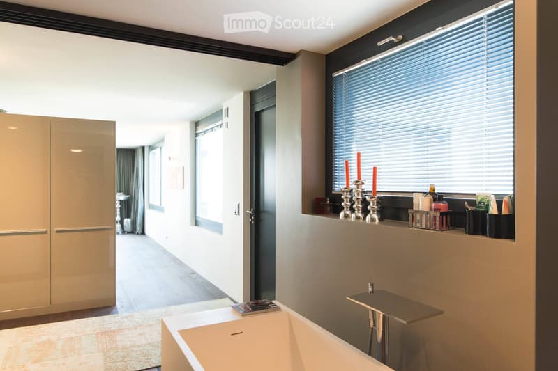 Modern bathroom with a freestanding bathtub, a small table, candle holders on the windowsill, a large mirrored wardrobe, and a towel rack. The space features large windows with blinds and a sleek, contemporary design.