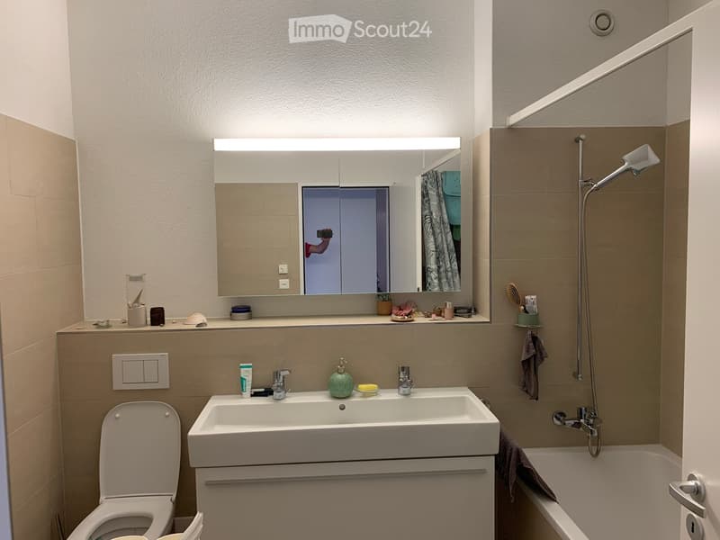 Modern bathroom with a white sink, toilet, bathtub, and a large mirror with built-in lighting. The space features beige tiles and a sleek, contemporary design.