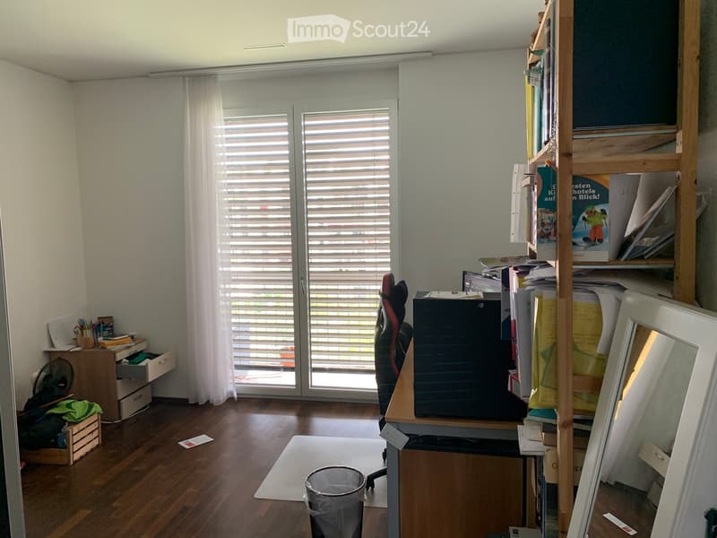 A well-lit room with wooden flooring, large windows with blinds, a desk with a laptop, a chair, a shelf with books, and a fan. The room also includes a small table with office supplies and a mirror on the wall.