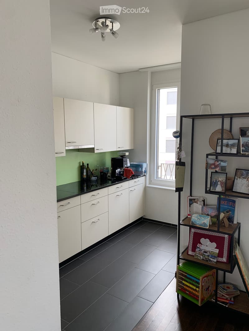 Modern kitchen with white cabinets, dark tiled floor, and a window overlooking a balcony. Includes a coffee maker, fridge, and a shelf with books and framed photos.