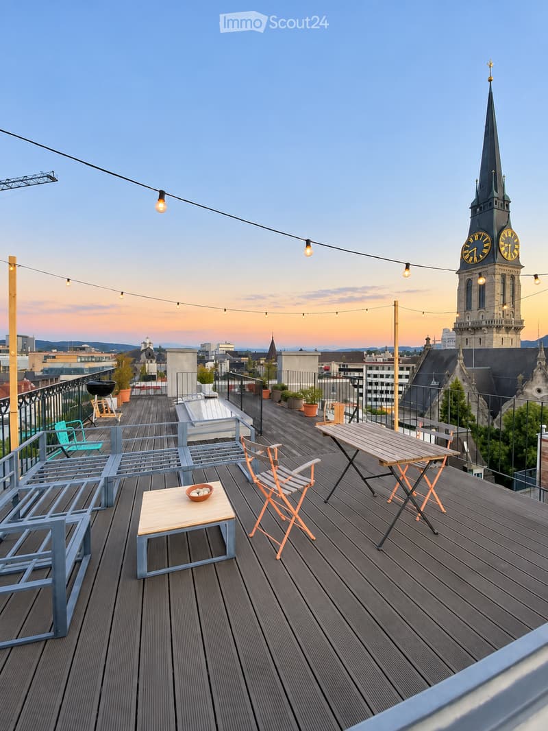 Rooftop terrace with wooden decking, string lights, a small table with a bowl, chairs, a bench, and a barbecue, overlooking a cityscape with a prominent church tower.