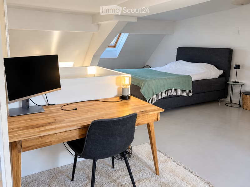 Modern bedroom with a large bed, desk with a screen, chair, and a small side table with a lamp. The room features a skylight window and a cozy green throw on the bed.