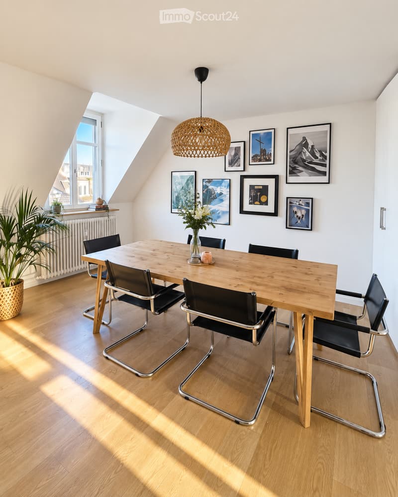 Bright, airy dining area with a wooden dining table surrounded by black chairs, a pendant light, and framed artwork on the walls. A potted plant and radiator are visible near the window.