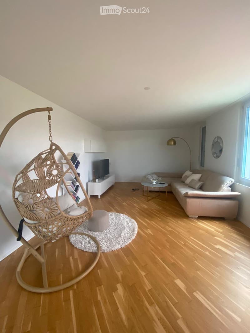 Modern living room with light wooden flooring, white walls, a beige sofa, a wicker hanging chair, a coffee table, and a TV stand.