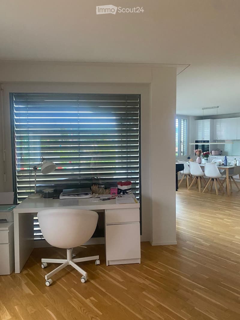 A modern, well-lit room with a desk, chair, and lamp on the left, and a dining area with a table and chairs on the right. The room features wooden flooring and large windows with blinds.