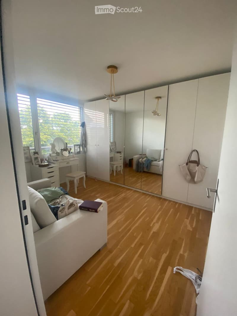 A well-lit room with wooden flooring, a white sofa, a large mirrored wardrobe, a small table with a chair, and a vanity area with a mirror and stool.