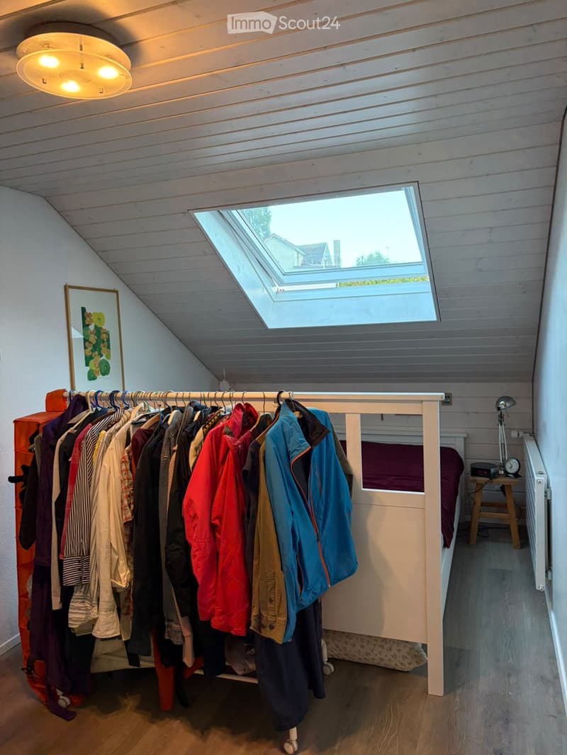 A small, cozy attic room with wooden walls and ceiling, featuring a lofted bed, a clothing rack, a picture frame, and a small table with a lamp.