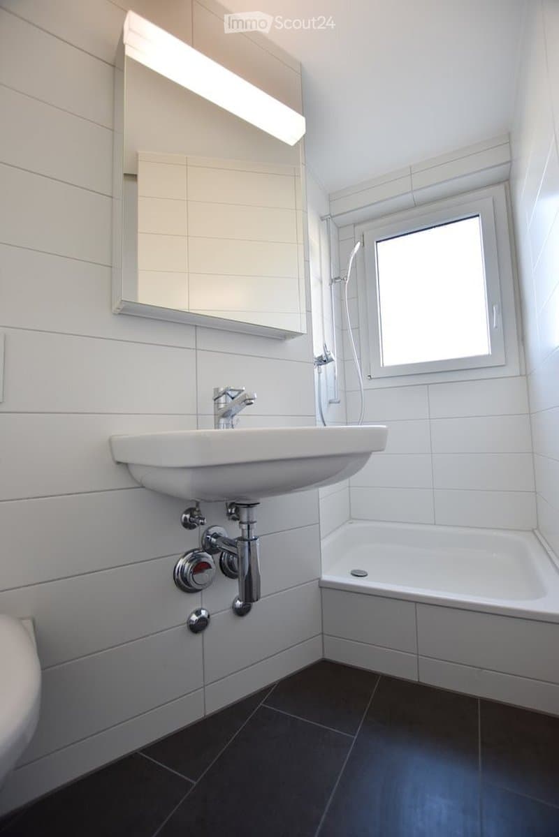 Modern bathroom with white tiled walls, a white sink with a chrome faucet, a bathtub, a mirror with a built-in light fixture, and a window providing natural light.