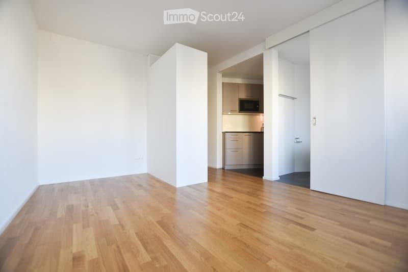 Empty room with wooden flooring and white walls, featuring an open doorway to a small kitchen area with cabinets and a microwave.