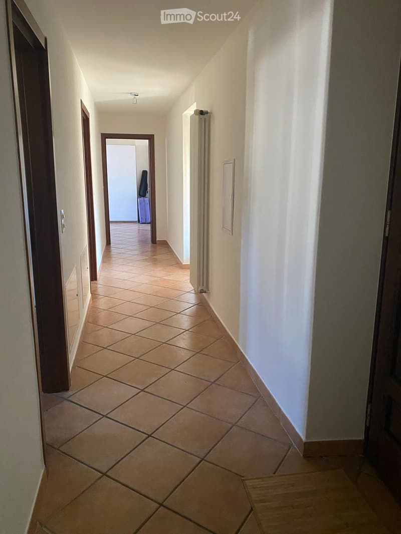 A narrow hallway with tiled flooring, white walls, and several doors leading to other rooms. A radiator is visible on one wall, and a small piece of furniture is seen in an adjacent room.