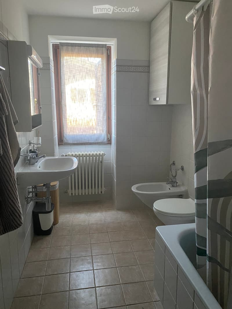 Bathroom with a sink, toilet, bathtub, and radiator. Features tiled walls and floor, a window with blinds, and a cabinet above the sink.
