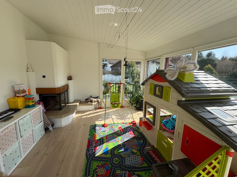 Brightly lit room with wooden flooring, fireplace, large windows, toy house, swing, storage unit, and colorful play mat.