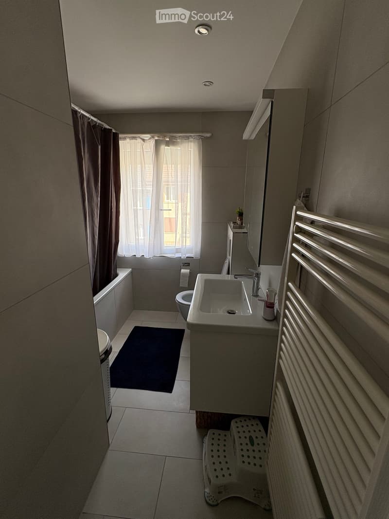 Modern bathroom with a sink, toilet, bathtub, radiator, and a small stool near the sink. The room features a window with sheer curtains and a dark bathmat on the floor.